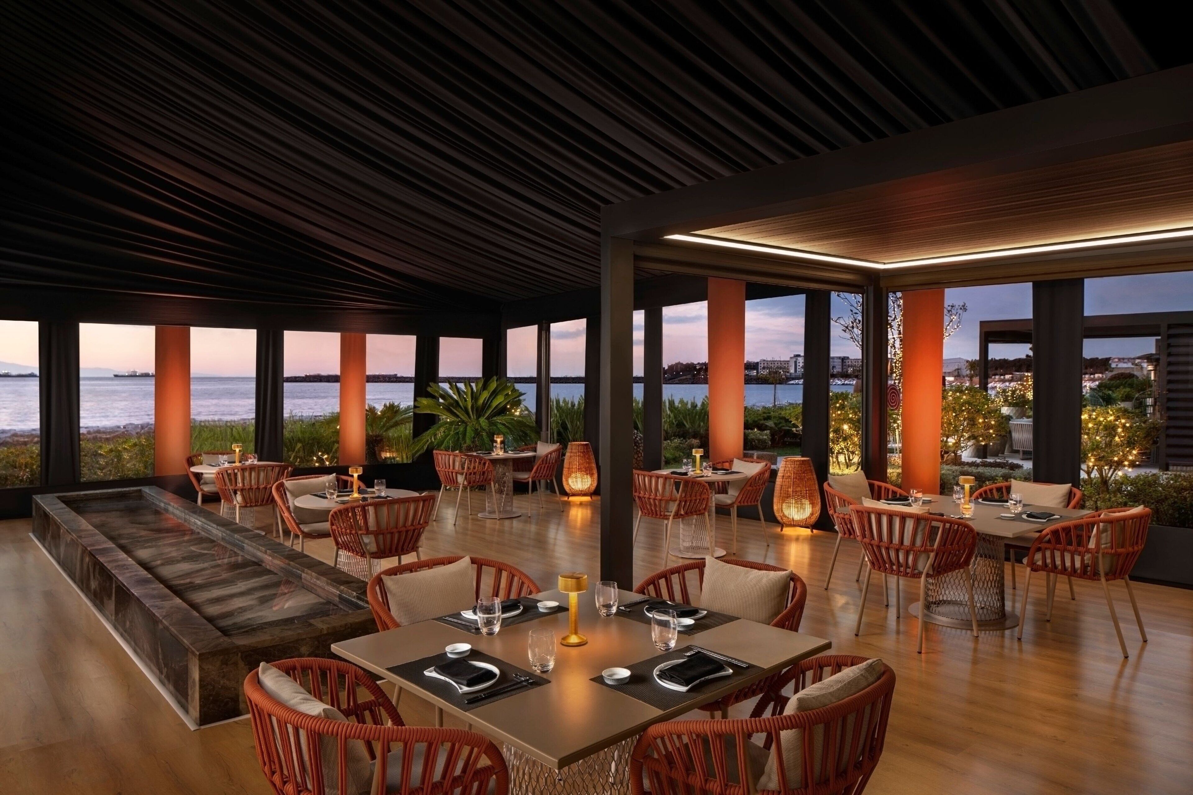 A spacious outdoor dining area with multiple tables and chairs, large windows offering a view of the sea, and a modern architectural design with a sloped ceiling.