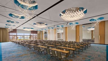 A conference room with rows of tables and chairs, large chandeliers, and a view of the outdoors.