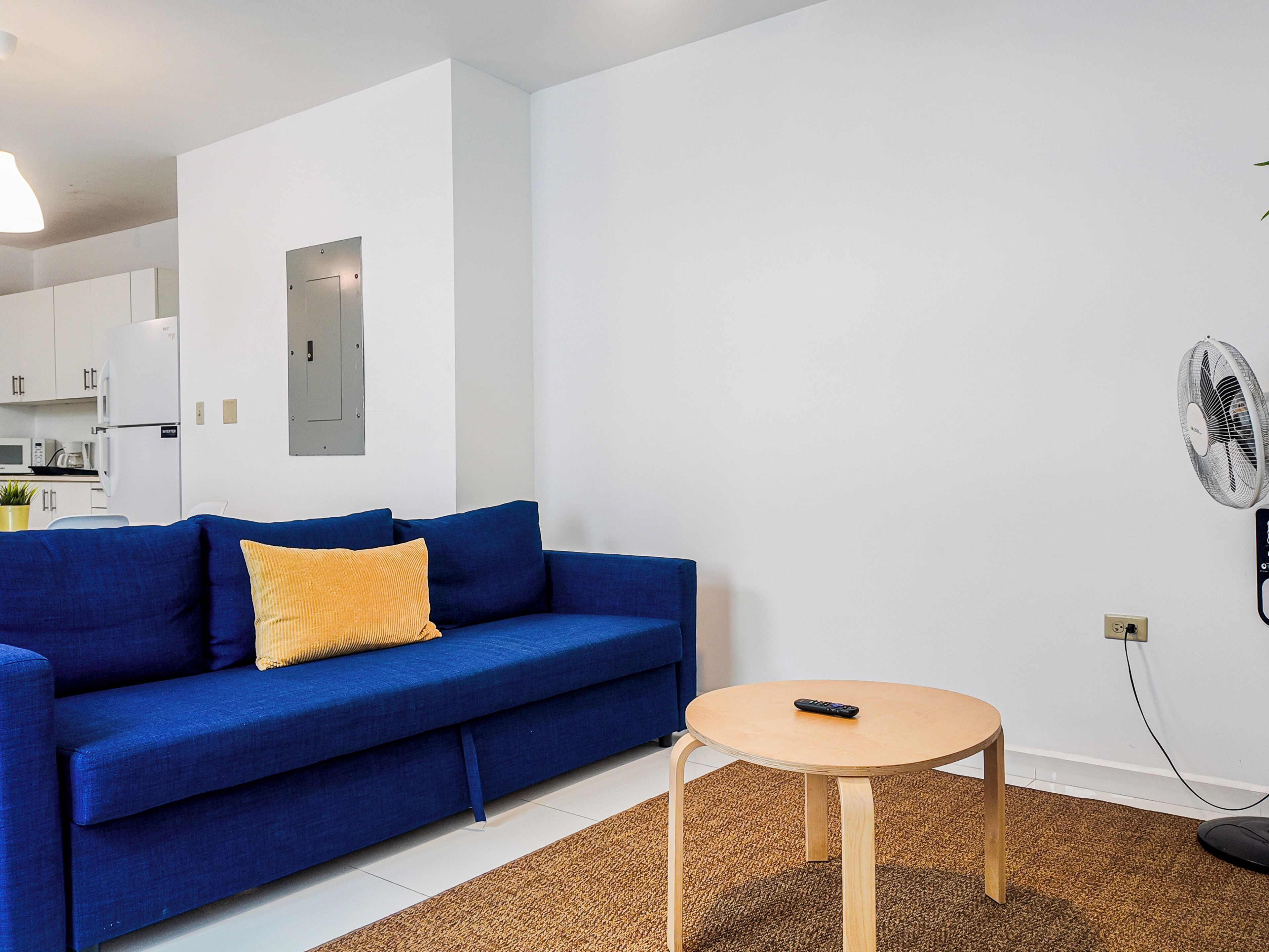 Uma sala de estar moderna com um sofá azul, uma mesinha de madeira e uma parede branca com porta e ventilador fixado.