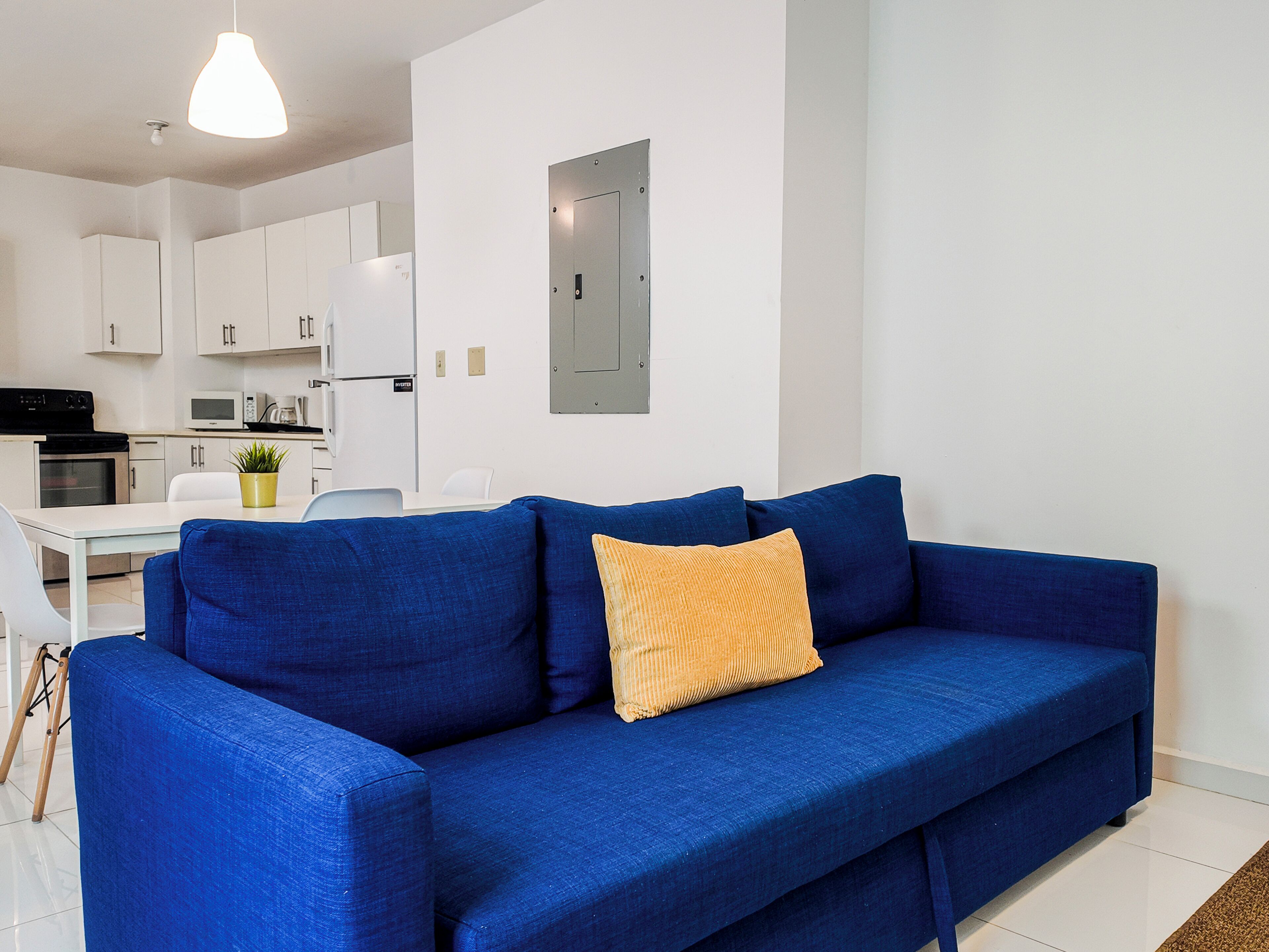 Una sala de estar moderna con un sofá azul, un cojín amarillo y una cocina al fondo.