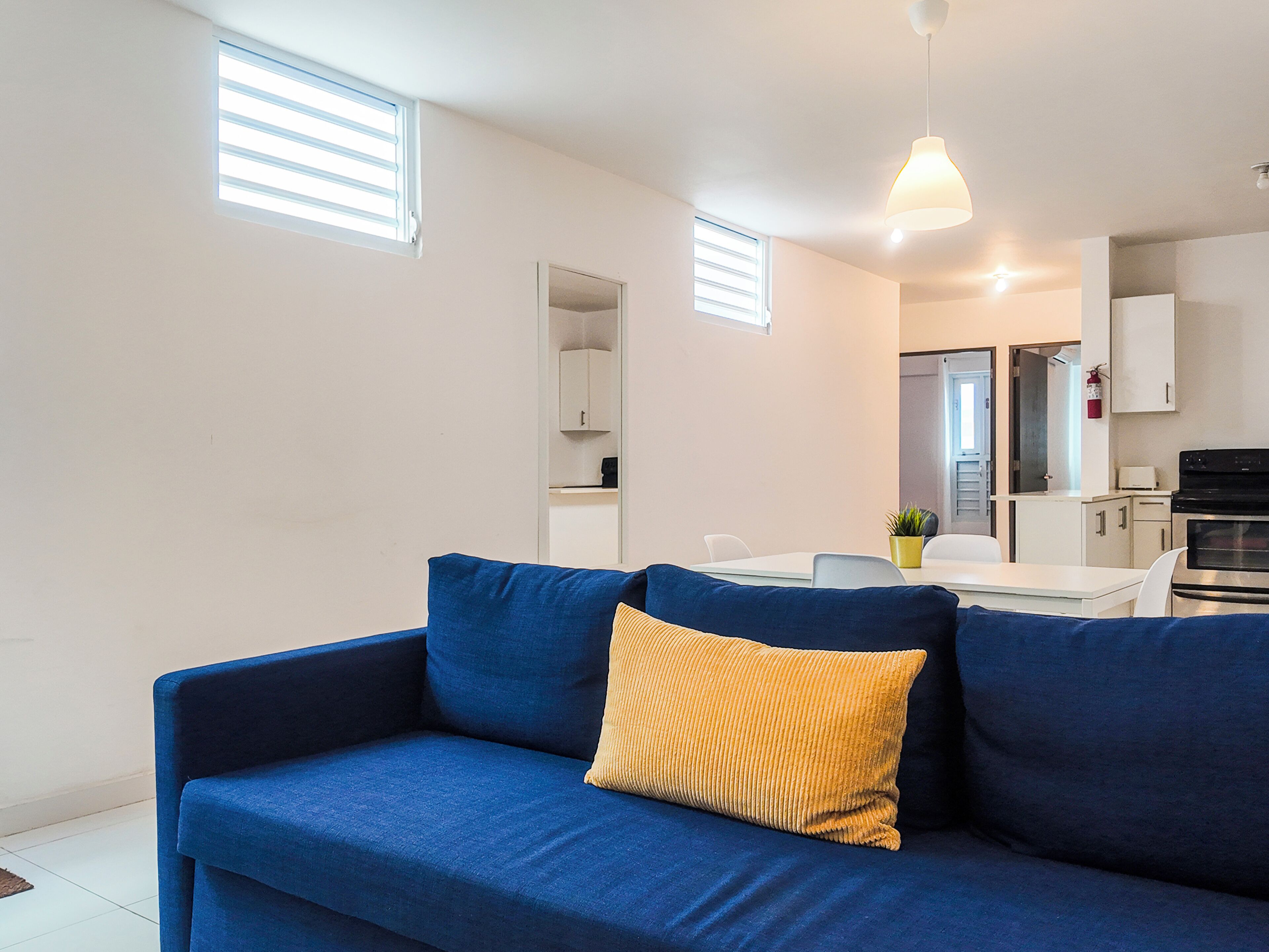 Een moderne woonkamer met een blauwe bank, een geel kussen, een eettafel en een keuken op de achtergrond.