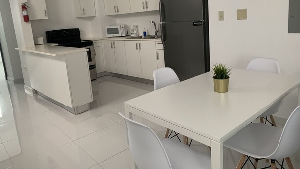 Eine moderne Küche mit weißen Schränken, einem schwarzen Kühlschrank, einer Mikrowelle und einer Kaffeemaschine. Ein Esstisch mit Stühlen und ein kleiner Topf mit Pflanze darauf stehen bereit.
