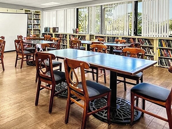 Uma biblioteca com mesas e cadeiras de madeira, estantes e grandes janelas.