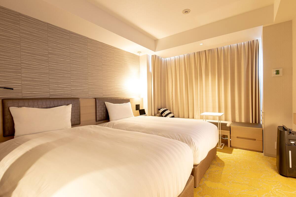 A hotel room with two large beds, a bedside table, a lamp, a wardrobe, and a minibar.