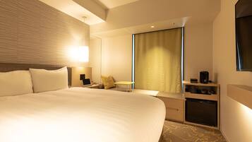 A hotel room with a large bed, bedside tables, a desk, a chair, a small kitchenette, and a large window with curtains.
