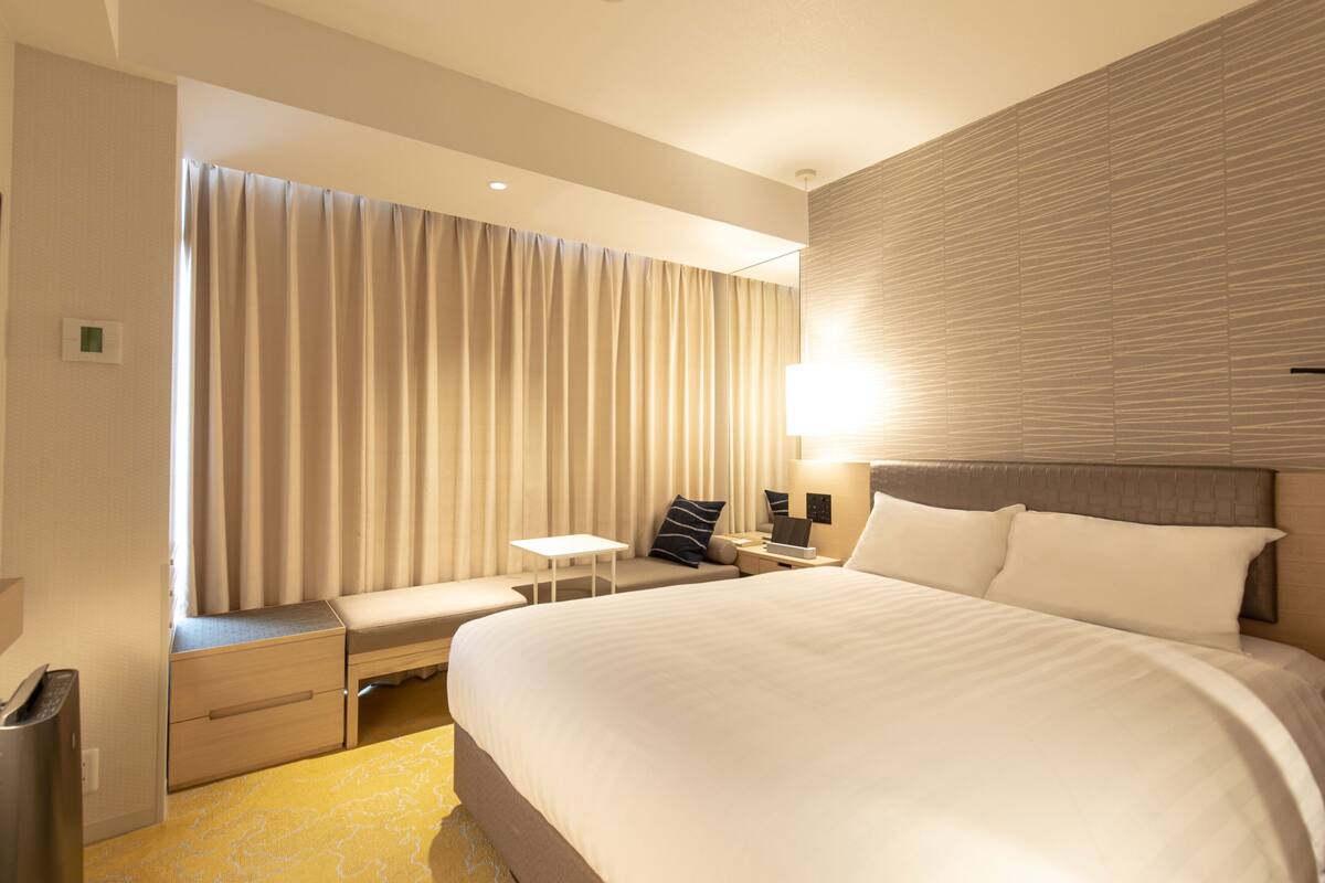 A hotel room with a large bed, a bedside table, a lamp, a bench, and a window with curtains.