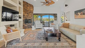 A spacious living room with a stone accent wall, a ceiling fan, and a large window offering a view of palm trees. - ★ Jump in! ★ Luxurious spread in the desert & no annoying neighbors! Comfy beds! (Fountain Hills)