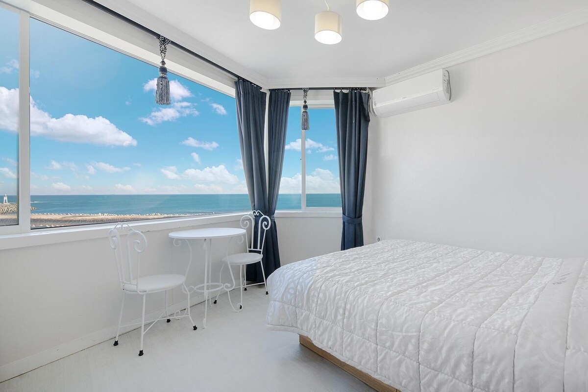 A bedroom with a bed, a small table with chairs, a window with curtains, and a view of the sea.