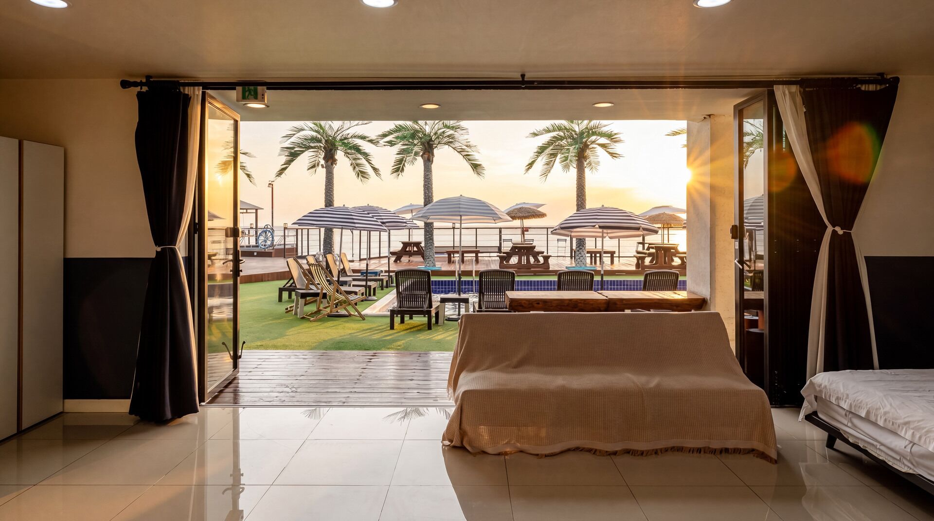 A hotel room with a bed, a view of an outdoor pool area with umbrellas, and a beach.