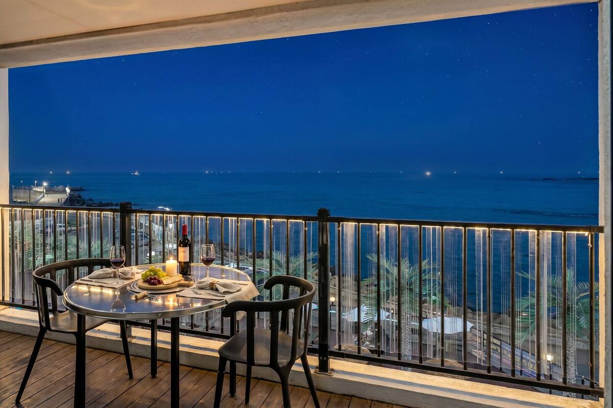 A balcony with a table set for two, overlooking a coastal view at night.
