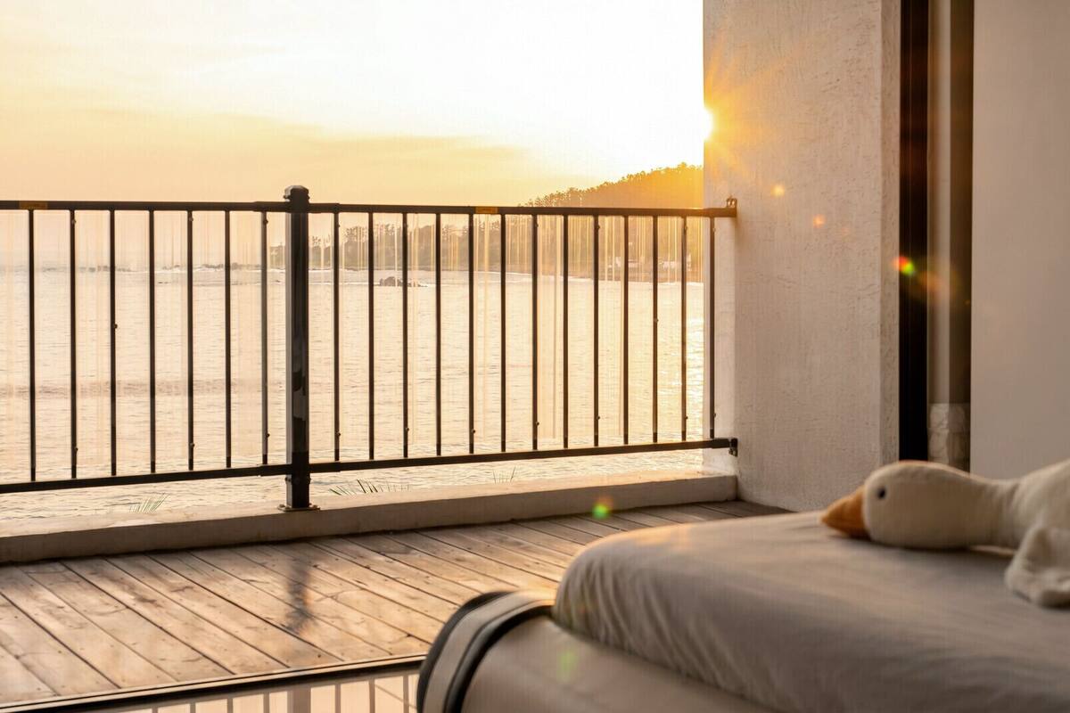 A balcony with a view of the sea, a bed with a pillow, and a wall with a window.