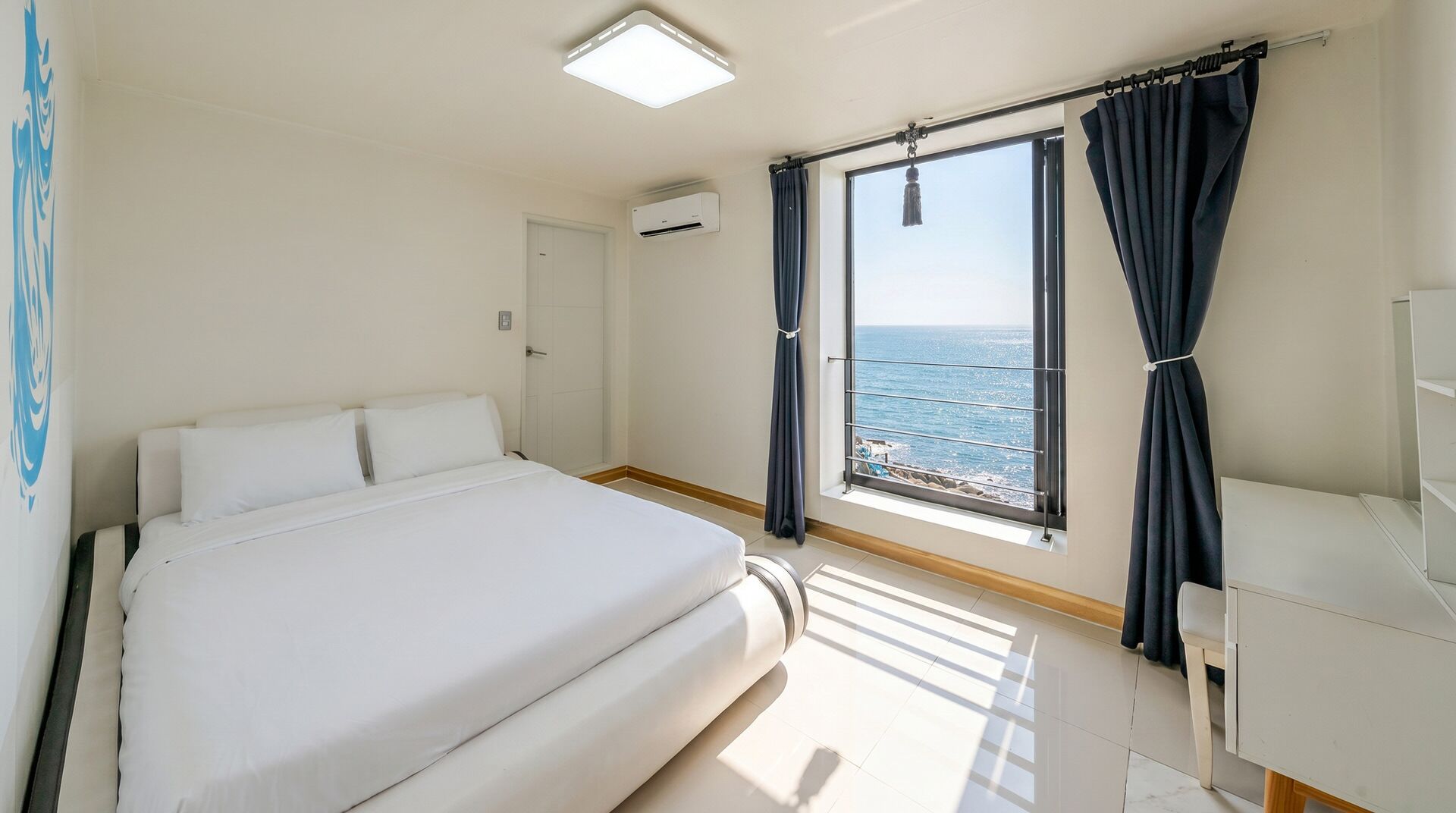A hotel room with a bed, a window with curtains, an air conditioning unit, and a view of the sea.