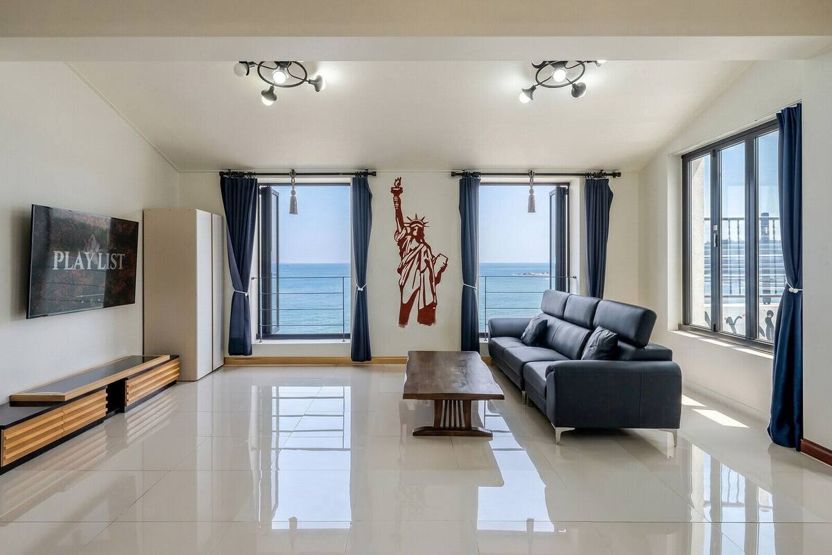 A modern living room with a large TV, a sofa, a coffee table, and a view of the sea.