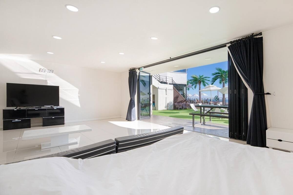 A modern bedroom with a large bed, a TV, and a balcony with a view of palm trees and a pool.
