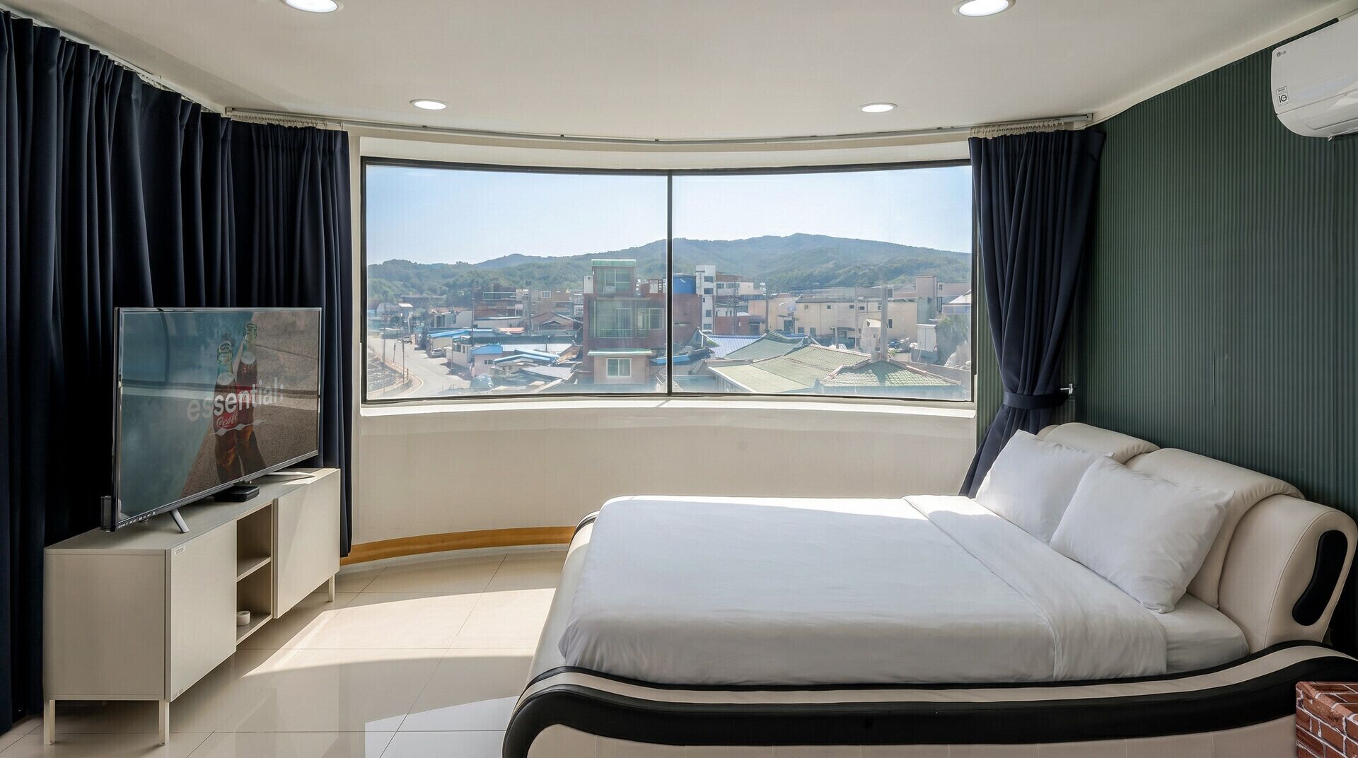 A hotel room with a large bed, a TV on a stand, and a view of a cityscape through a large window.