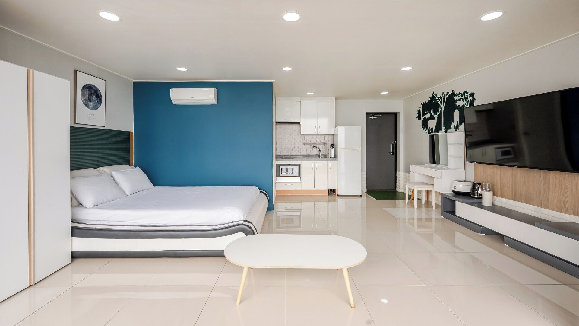 A modern hotel room with a bed, a small white table, a kitchenette, and a TV.