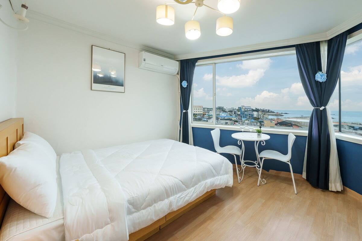 A hotel room with a bed, a small table with chairs, a view of the city and sea, and a ceiling light fixture.