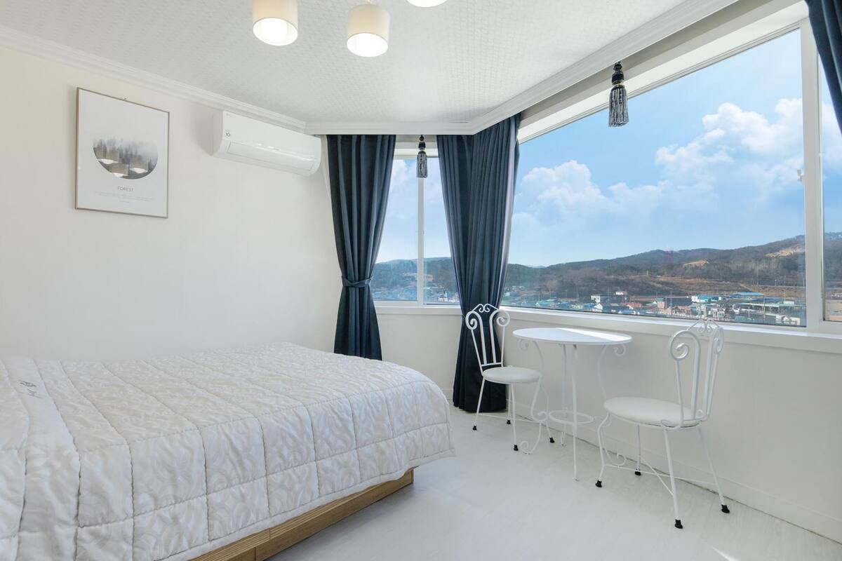 A bedroom with a bed, a small table, two chairs, a window with a view of the city, and a framed picture on the wall.