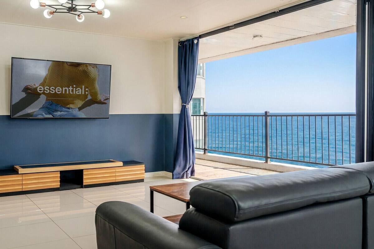 A modern living room with a large TV, a sofa, a coffee table, and a view of the sea.