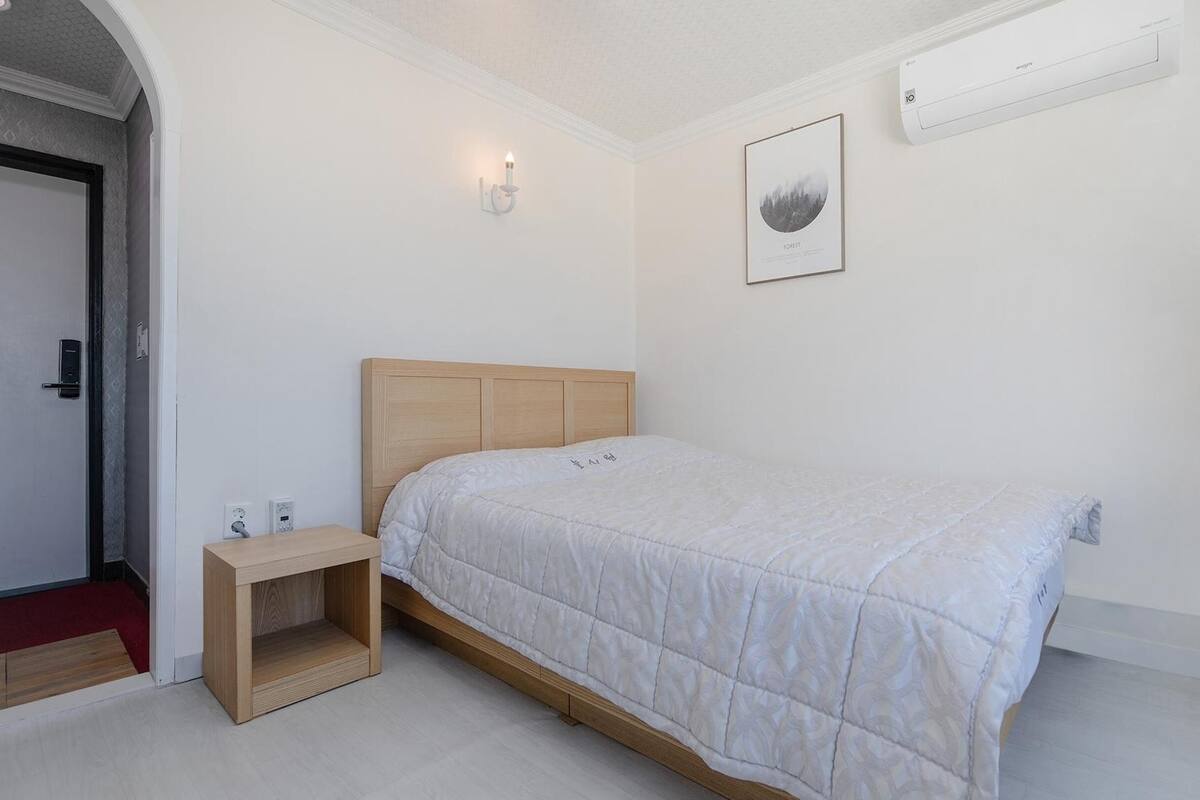 A bedroom with a bed, bedside table, wall-mounted air conditioner, and a framed picture.