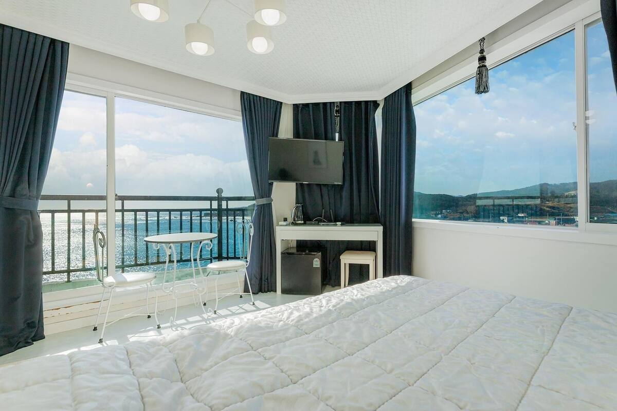 A bedroom with a large bed, a TV, a desk, and a balcony with a view of the sea.