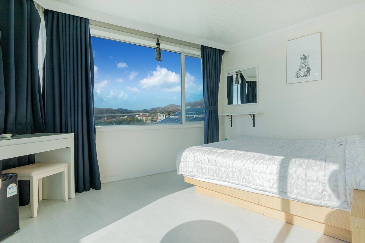 A bedroom with a bed, a desk, a mirror, and a window with a view of buildings and hills.