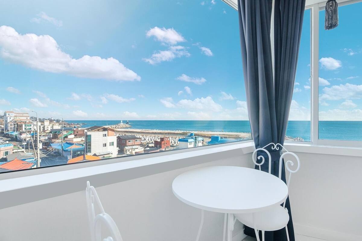 A balcony with a white table and chairs, overlooking a coastal town with buildings and a beach.