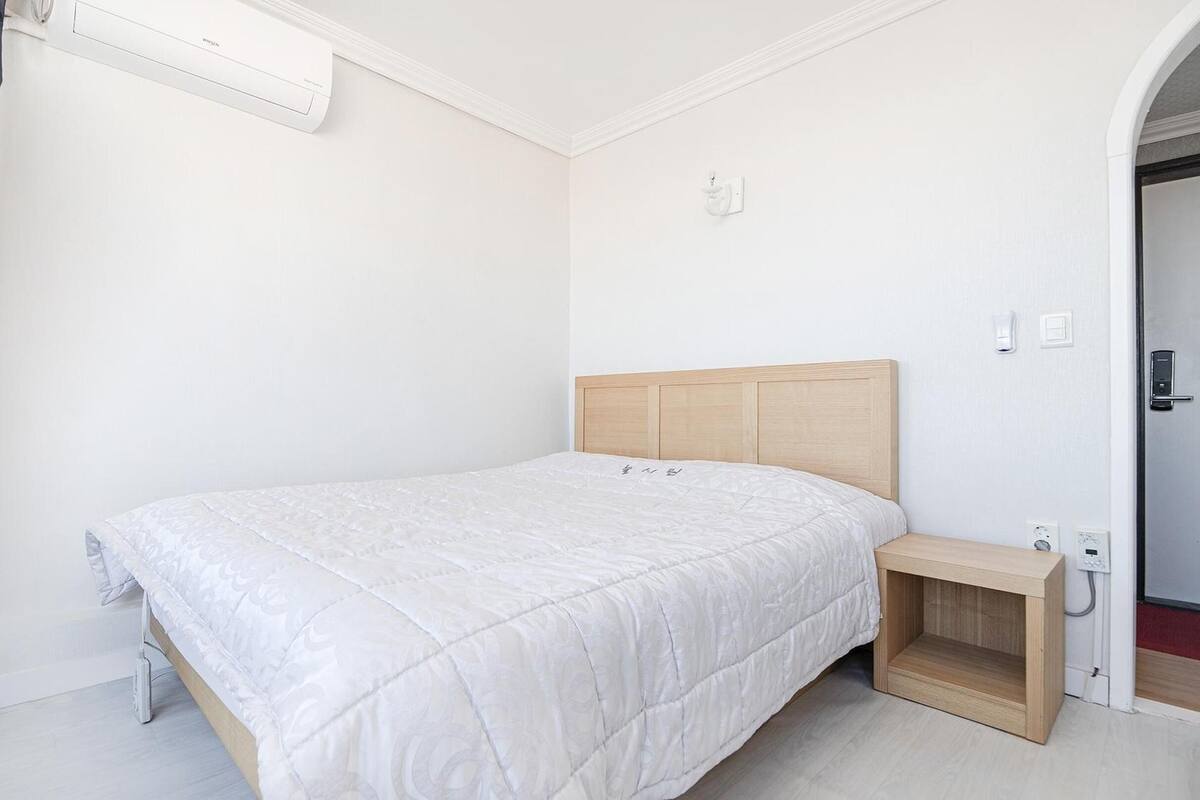 A bedroom with a bed, a bedside table, a wall-mounted air conditioner, and a door with a handle.