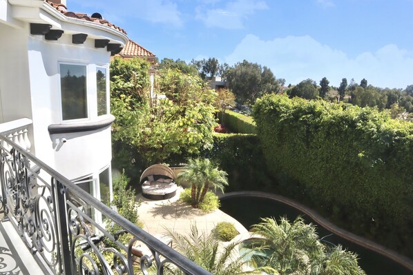 A balcony with a wrought iron railing, a white building with a tiled roof, and a well-maintained garden with a curved pathway and a hammock.