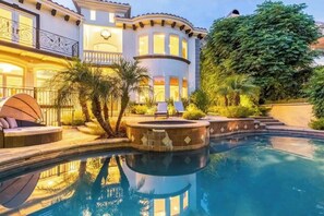 Ein luxuriöser Poolbereich mit einer gewölbten Poolanlage, einer Sitzecke mit Liege und einem großen Haus mit Balkon und beleuchteten Fenstern.