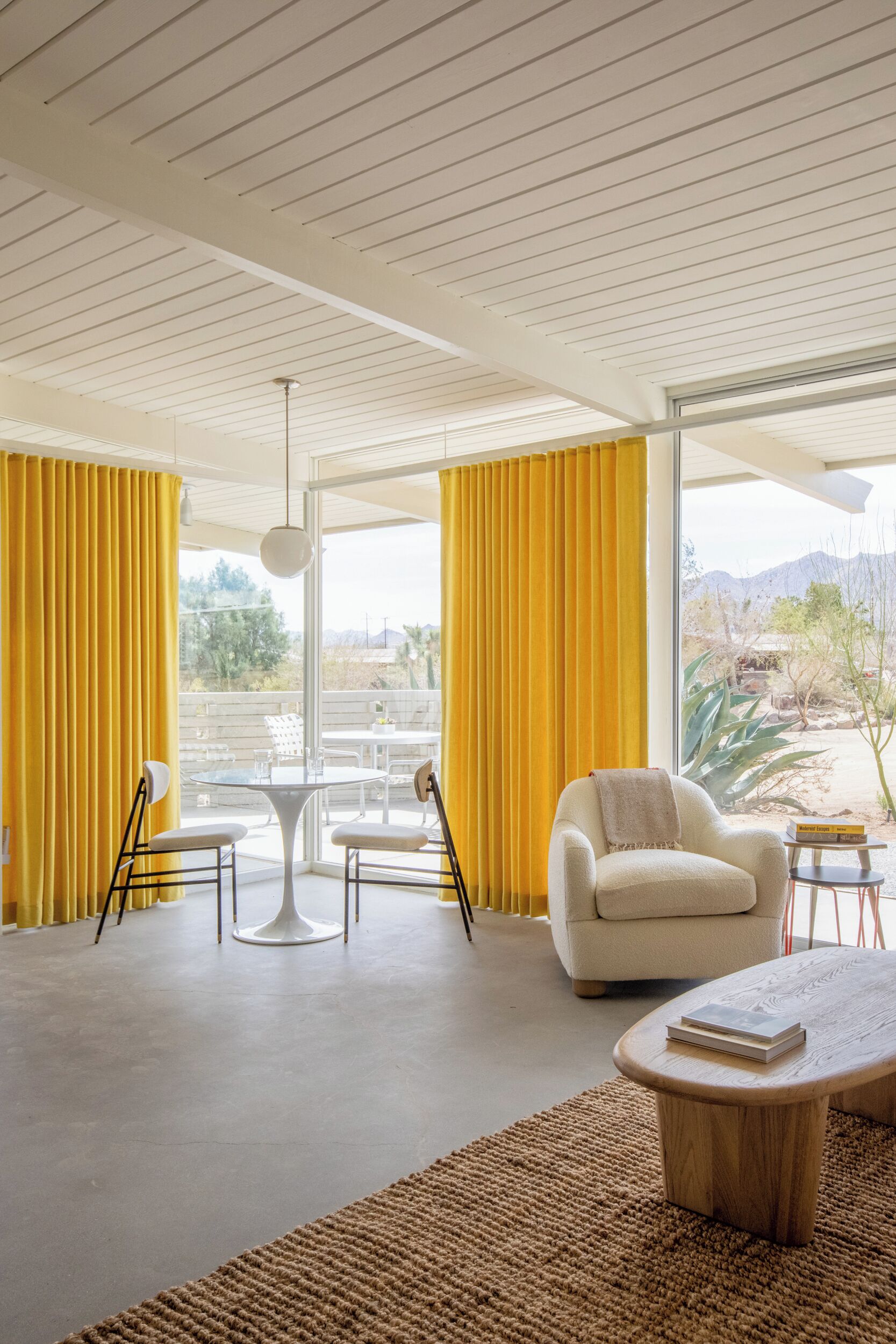 Uma sala de estar moderna com um sofá branco, uma mesa de centro e uma mesa de jantar com cadeiras. Grandes janelas com cortinas amarelas oferecem vista para o exterior.