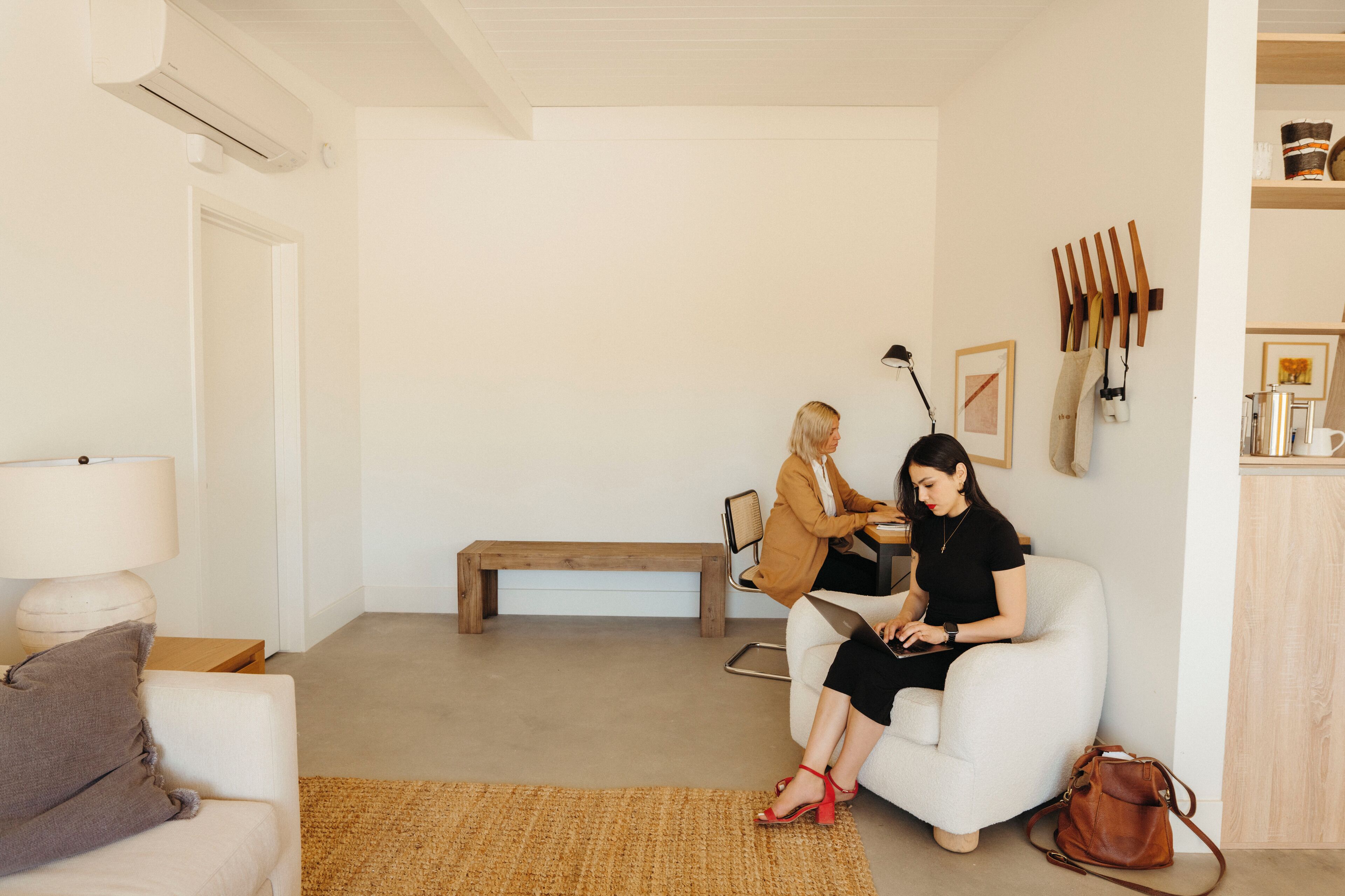 A modern living room with a white sofa, a wooden bench, and a coffee table. There is a lamp on the left side and a brown bag on the right.