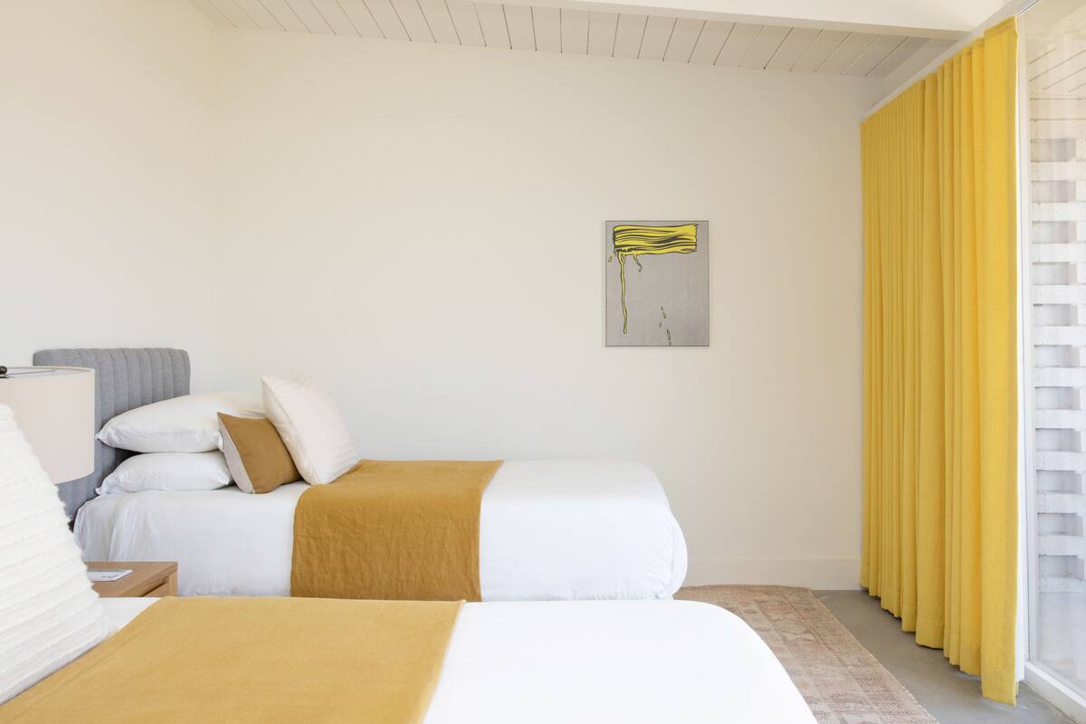 Ein Hotelzimmer mit zwei Betten, einem Fenster mit gelben Vorhängen und einem Bild an der Wand.