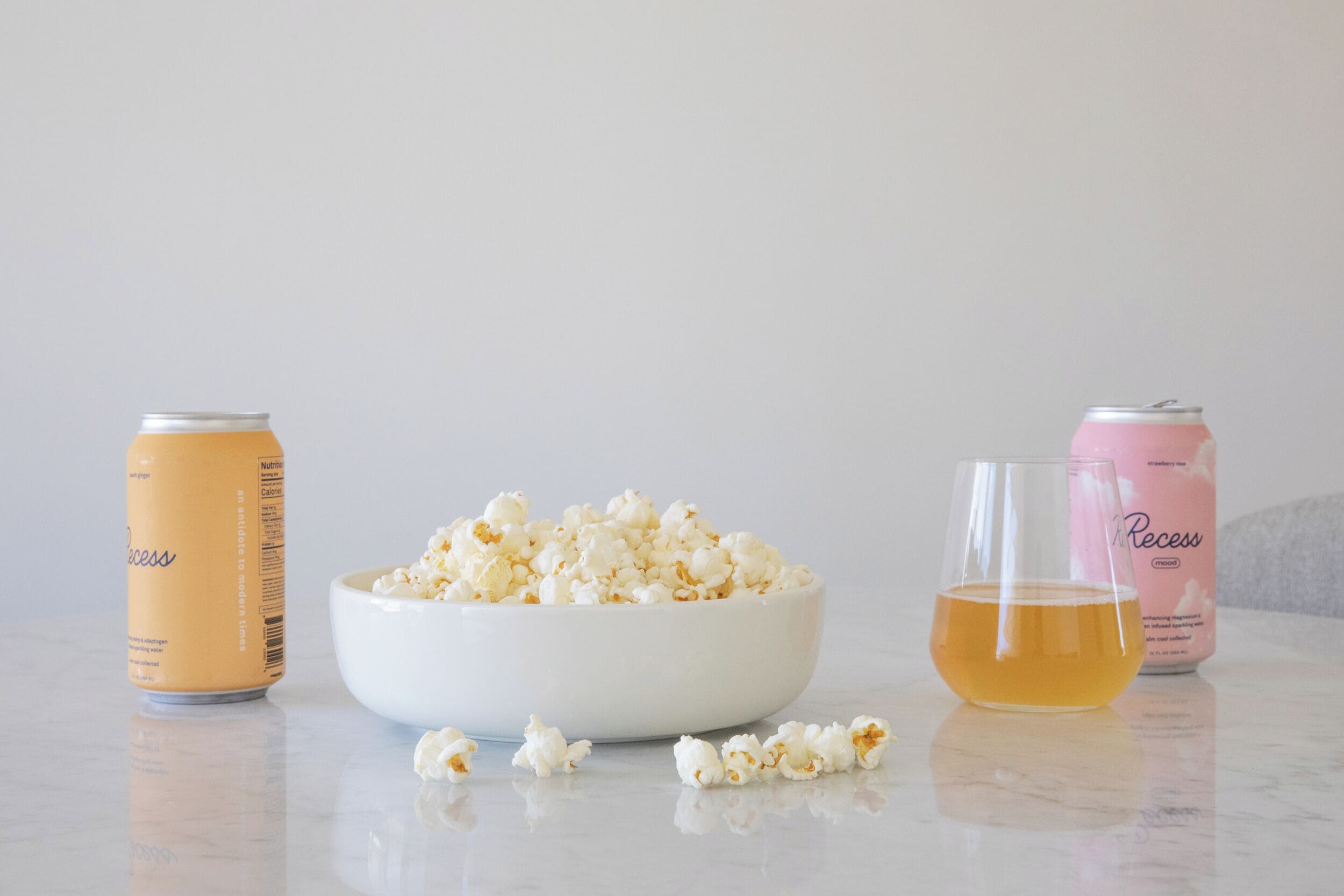 Popcorn i en skål, et glas gylden væske og to dåser drikke på en reflekterende overflade.