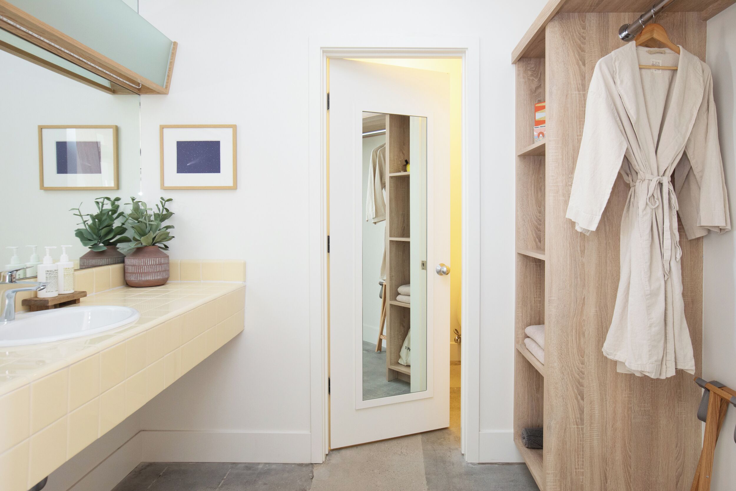 Un baño con lavamanos, espejo, cuadros enmarcados, planta en maceta y puerta de madera que da a un armario con una bata colgada.