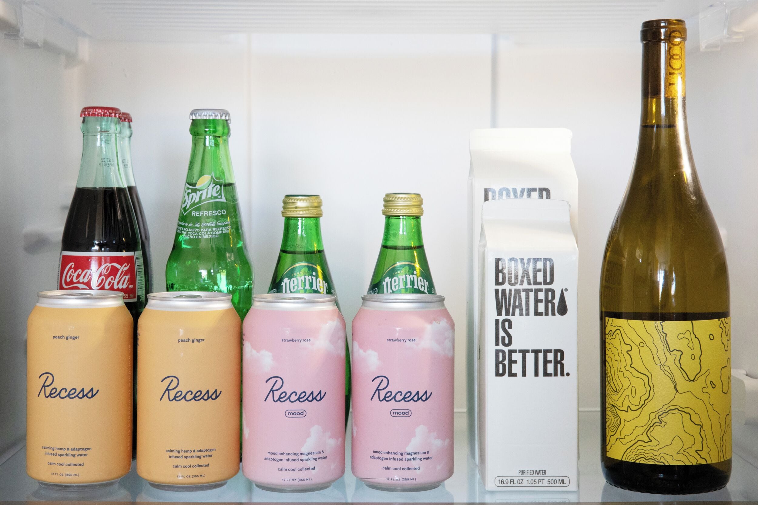 Une étagère de réfrigérateur contenant diverses boissons, notamment des canettes de soda « Recess » et « Terrier », une bouteille de vin et un emballage de « Boxed Water ».