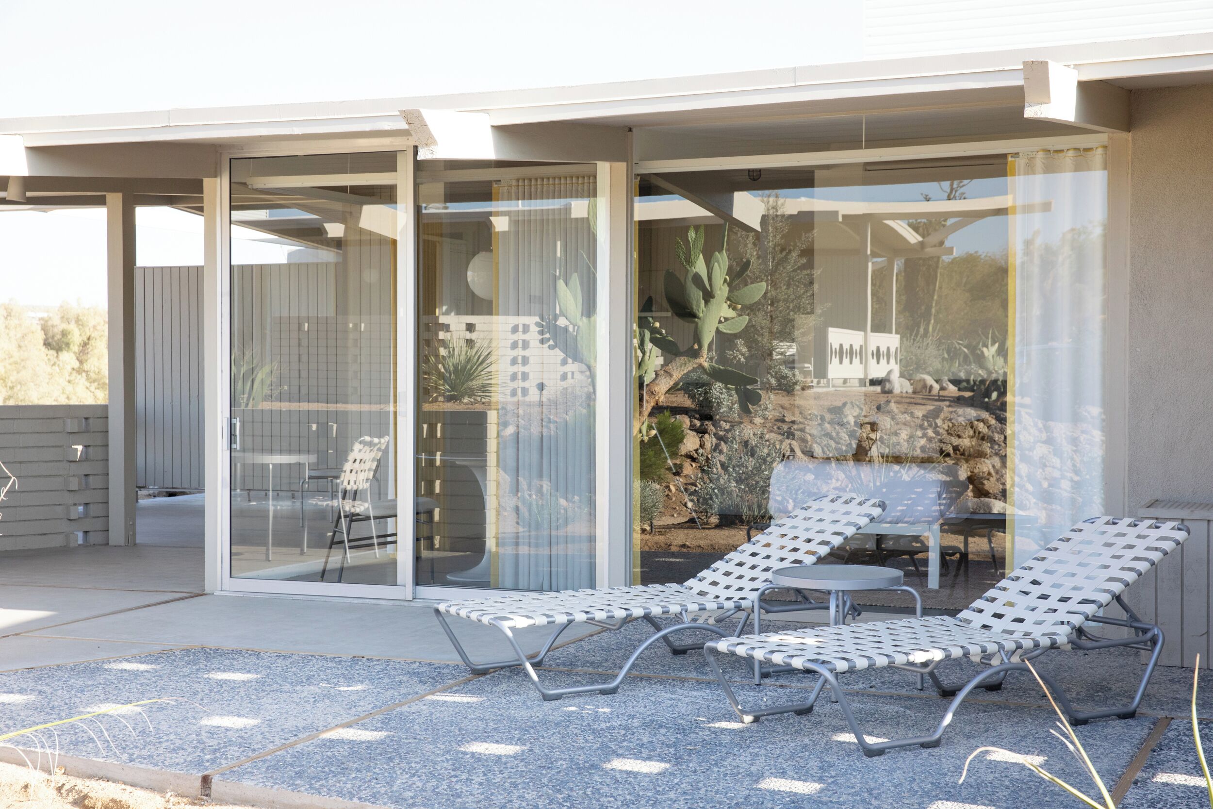 Un patio moderno al aire libre con tres sillones de descanso y una mesa, rodeado por una estructura de vidrio que ofrece vistas de plantas desérticas y un paisaje rocoso.