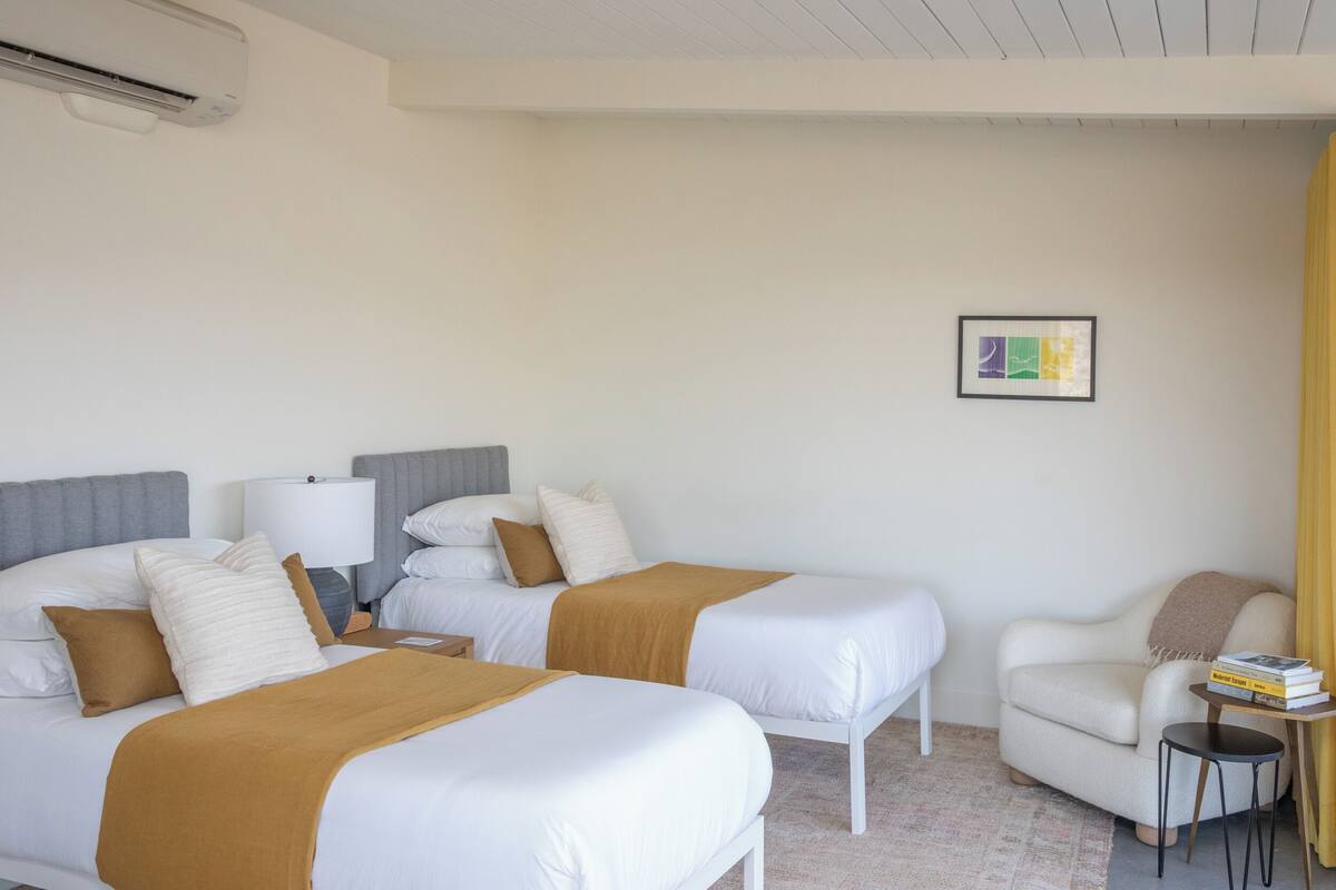 Ein Hotelzimmer mit zwei Betten, einem Sessel, einem kleinen Tisch mit Büchern und einer Wand-Klimaanlage.