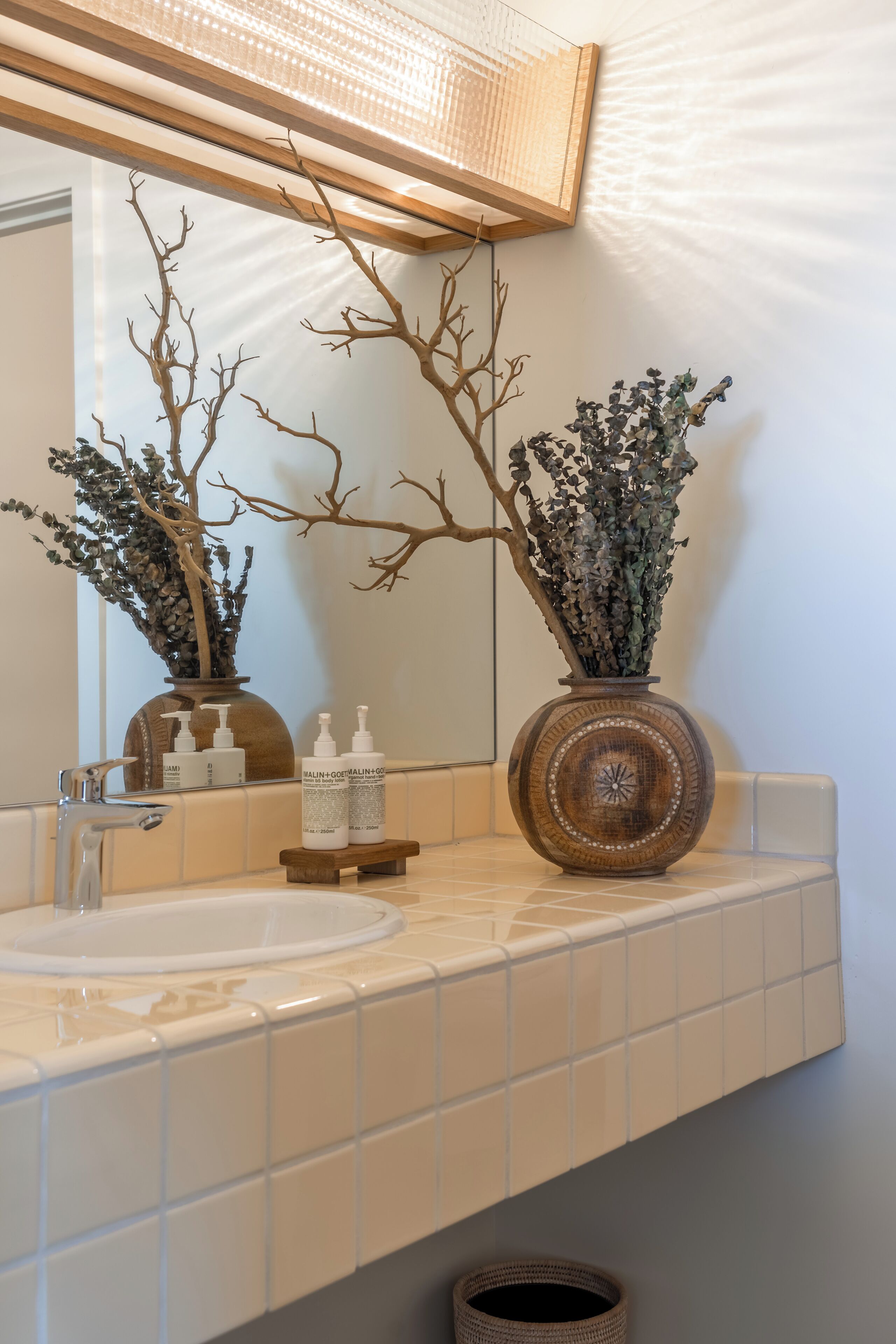 Un baño con un lavamanos de azulejos blancos, un espejo, dos jarrones decorativos y un estante de madera.