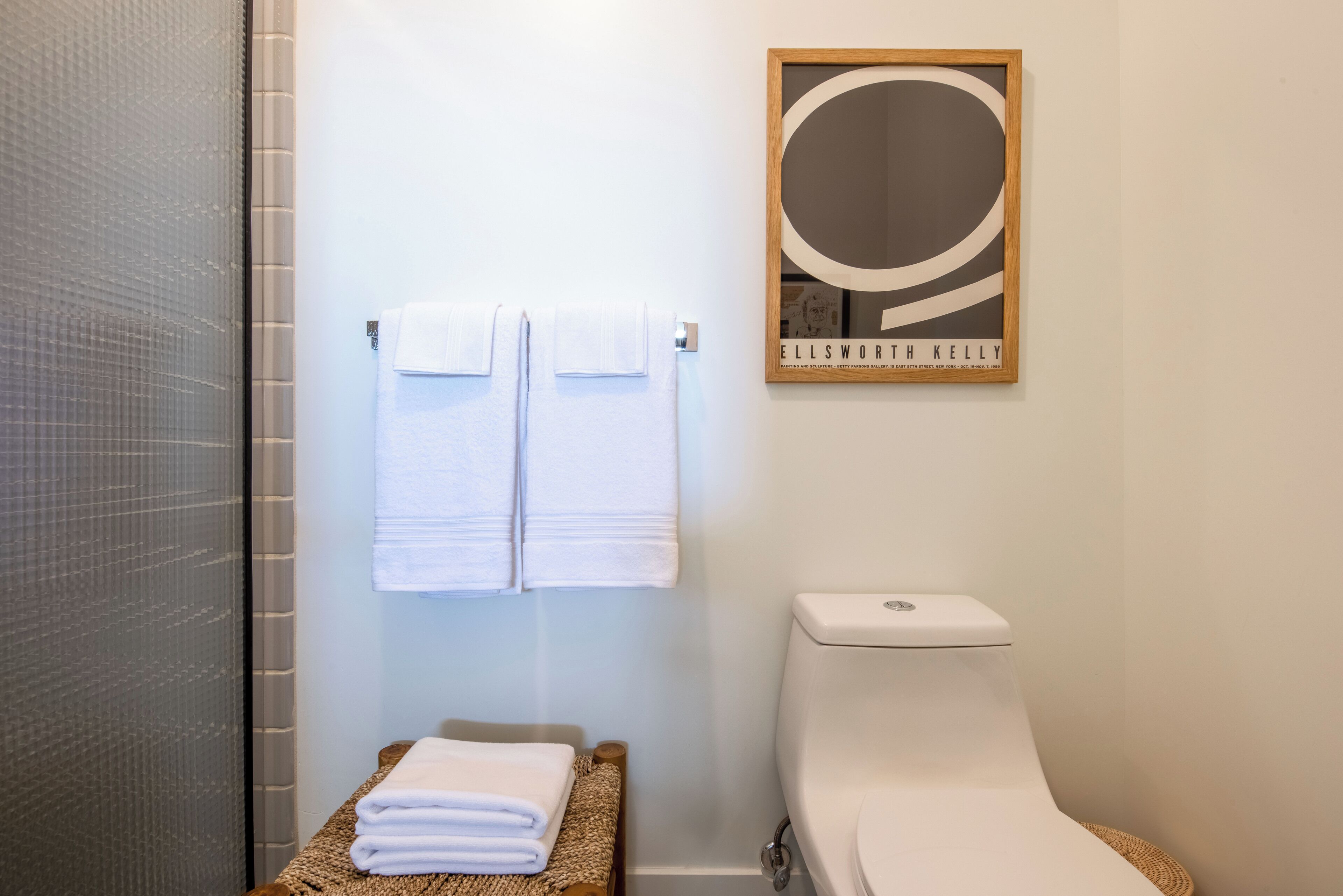 A bathroom with two white towels hanging on a rack, a framed artwork above the toilet, and a woven mat on the floor.