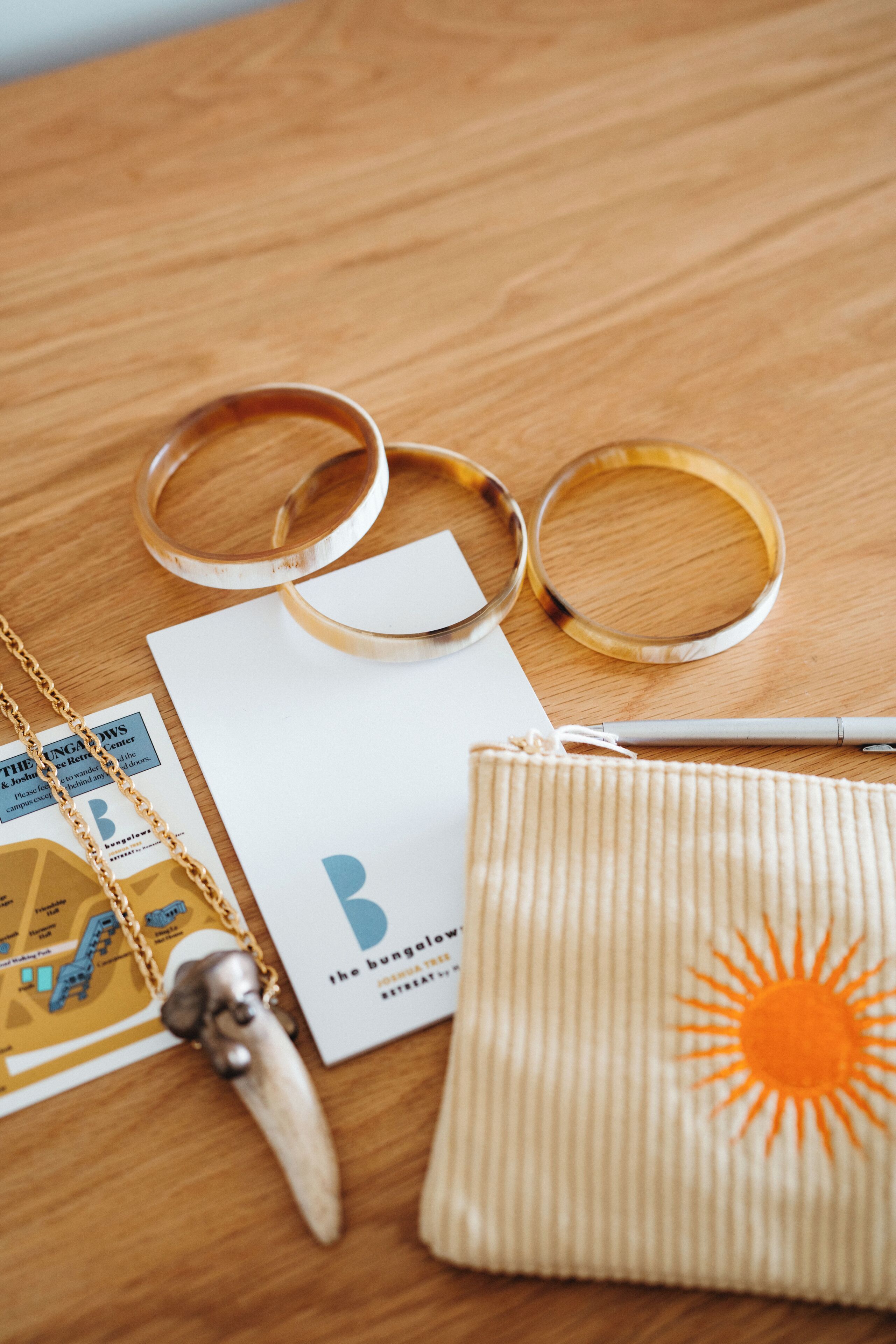 A wooden table with a white paper, a pen, a beige bag with a sun design, a wooden object, and two circular objects.