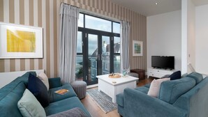 Una sala de estar moderna con papel tapiz a rayas, un ventanal con cortinas, un televisor de pantalla plana y una zona de estar acogedora con sofás y otomanas color turquesa.