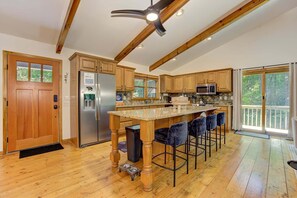 A spacious kitchen with wooden cabinets, a central island with bar stools, a stainless steel refrigerator, and a large window with a view of trees.