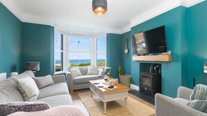 A living room with teal walls, a sofa, a coffee table, a fireplace, and a TV. - Porthmeor View, St Ives  – Sleeps 8 – On-Site Parking for Two Cars – Sea Views (St Ives)