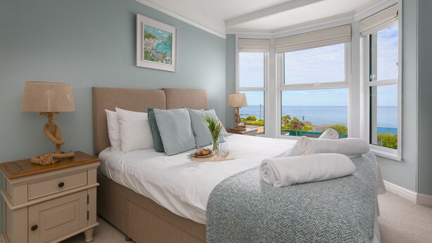 A bedroom with a large bed, a nightstand, a chair, a window with a view, and a painting on the wall. - Porthmeor View, St Ives  – Sleeps 8 – On-Site Parking for Two Cars – Sea Views (St Ives)