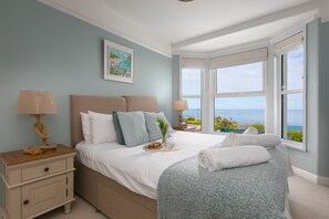 A bedroom with a large bed, a nightstand, a chair, a window with a view, and a painting on the wall.