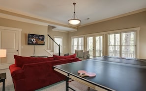 Una habitación con mesa de ping pong, un sofá rojo, televisor y ventanas amplias.