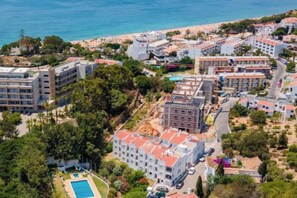 Vista aérea de uma área costeira com um hotel, vários edifícios, piscina e uma praia ao fundo.