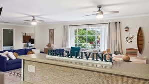 A modern living room with a central island, a large window with curtains, ceiling fans, and a 'BEACH LIVING' sign. - BIG Backyard w/Terrace|Dining|Games | 6min2Beach (St. Augustine)