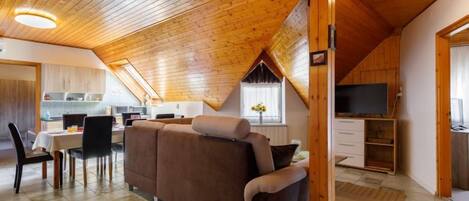 A cozy loft-style living area with a wooden ceiling, a sofa, a dining table, and a kitchen in the background.